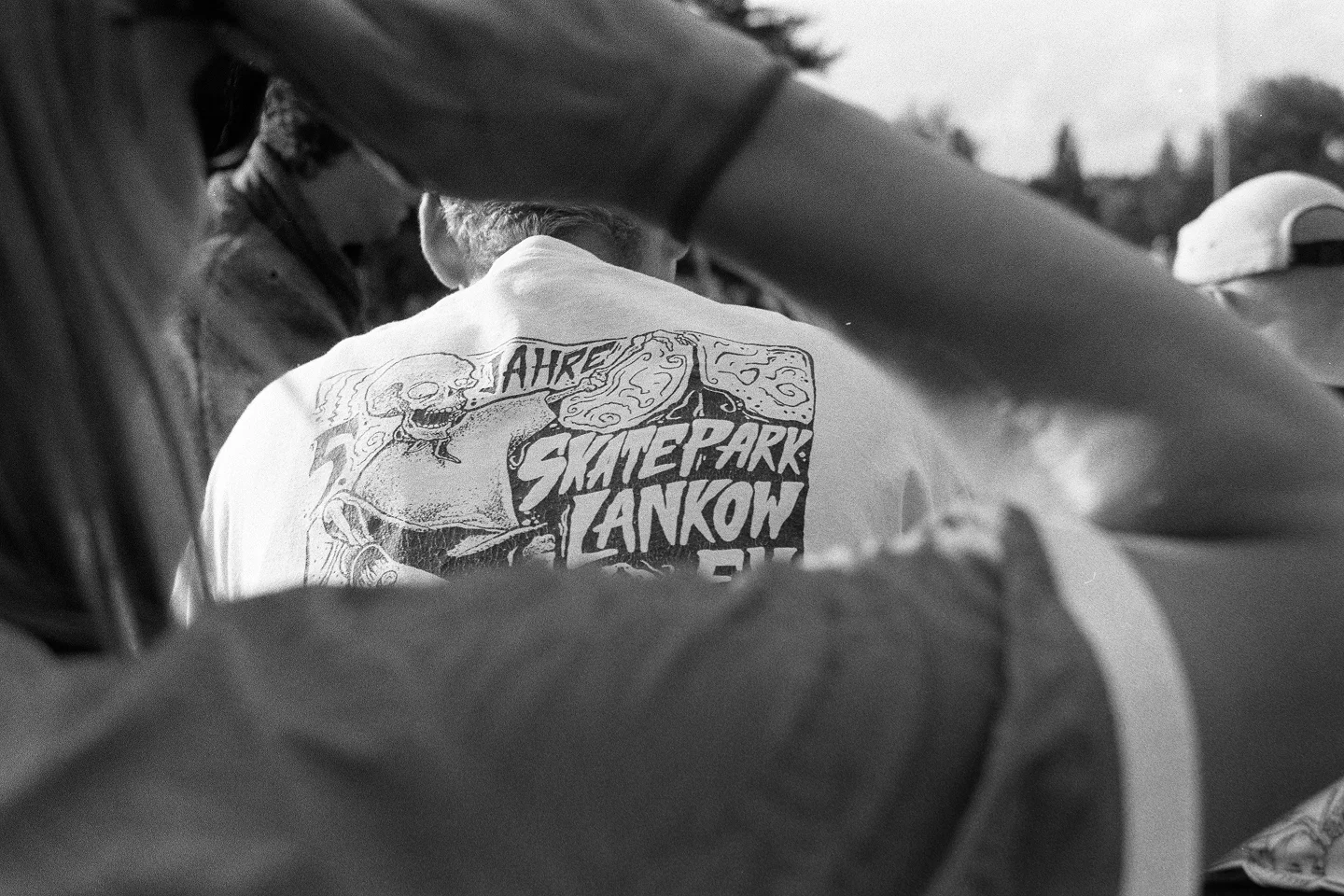 Back view of a person wearing a Skatepark Lankow shirt, partially framed by arms.