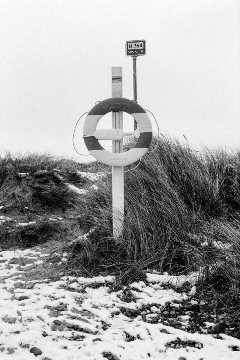 Life preserver ring mounted on emergency post with H 764 sign at snowy beach dunes surrounded by marram grass