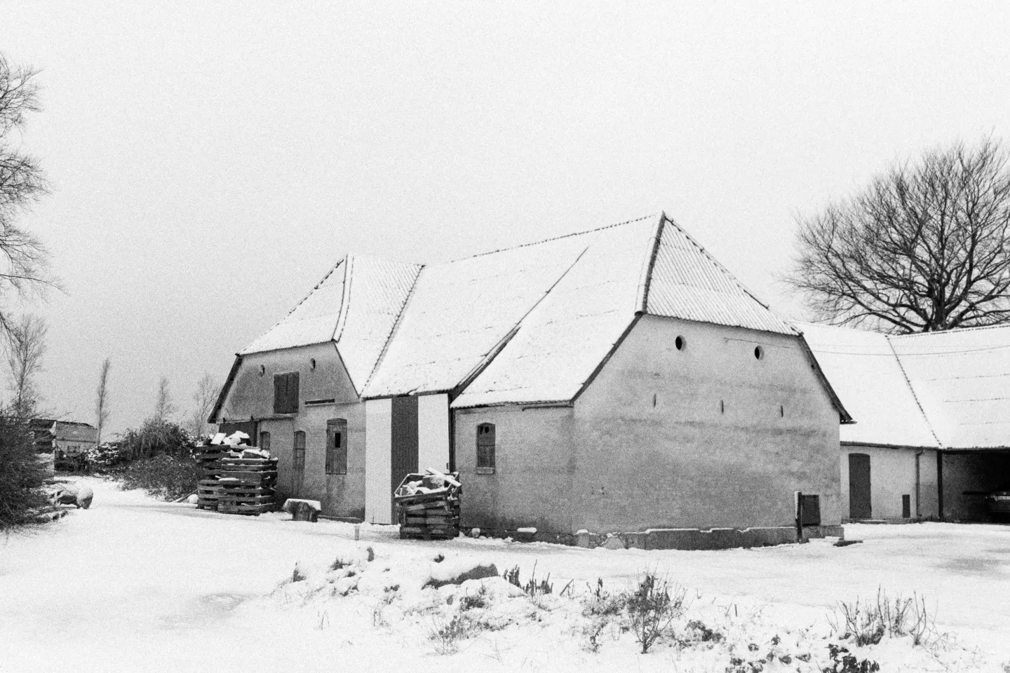 Traditional farm buildings with snow-covered roofs and stacked pallets in winter landscape