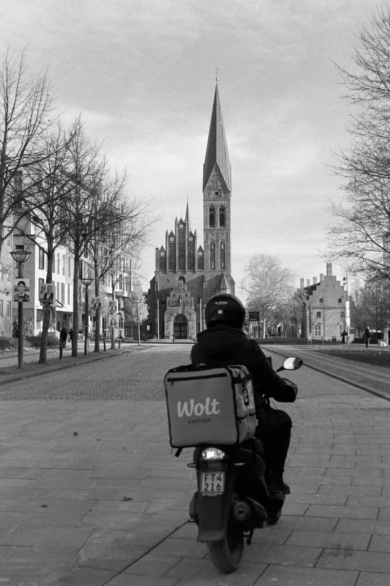 Wolt food delivery rider on scooter driving toward Gothic church in city square