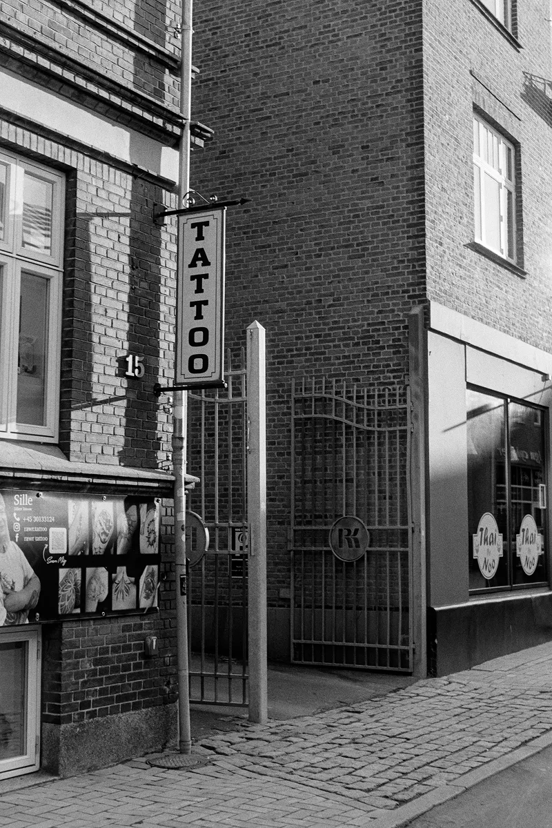 Tattoo studio entrance on cobblestone street between brick buildings with vertical sign