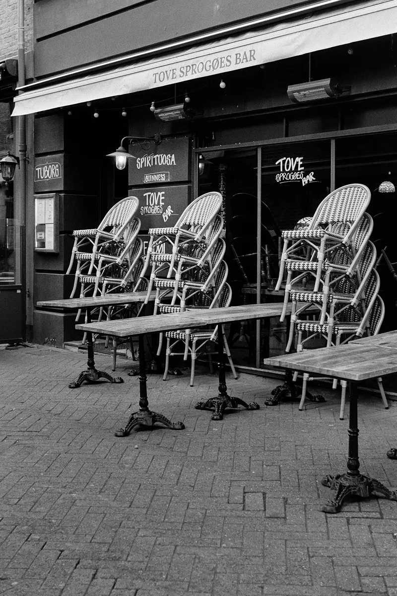 Closed outdoor terrace of Tove Sprogøes Bar with stacked bistro chairs and wooden tables