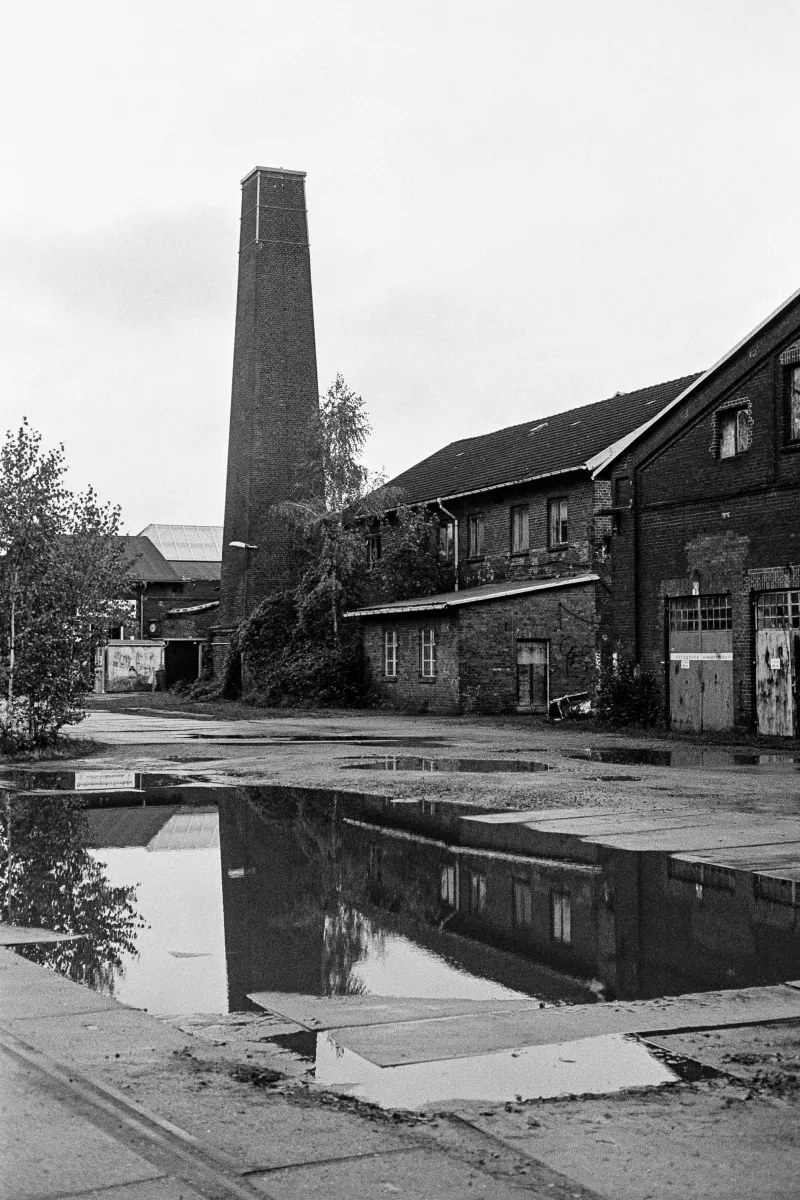 View of a large old building located behind the train station. A large square chimney is part of the scene and is reflected in a puddle in the foreground.