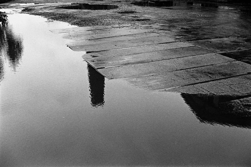 Detail of an old, rain-wet concrete slab roadway in rainy weather. In the foreground, a large puddle reflects a piece of a chimney.