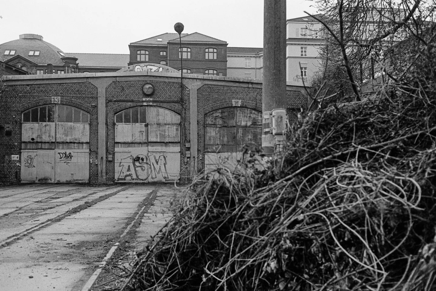 An old train station building that is no longer in use, with several large, locked gates leading to old tracks. Above it, a broken clock on the wall. In the foreground, a pile of brushwood.