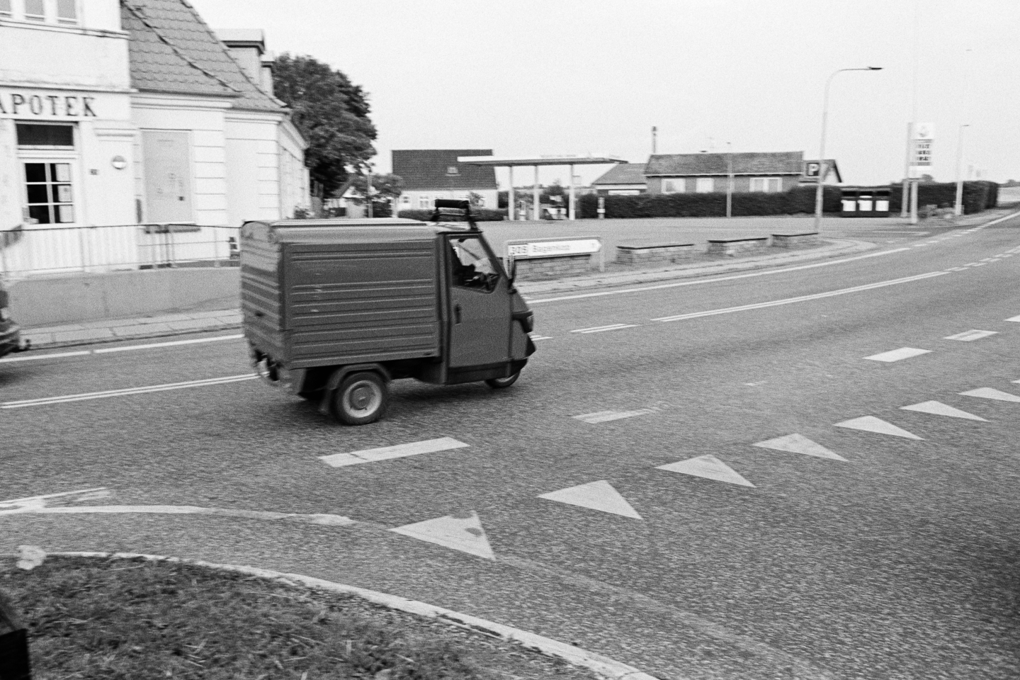 Three-wheeled delivery vehicle crossing zebra crossing on cobblestone street with pharmacy in background