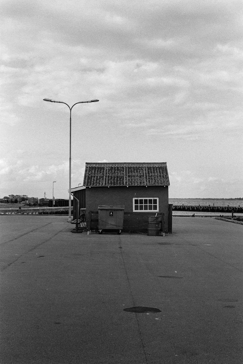 Symmetrical B&W film photo of a small shack and double street lamp on a desolate harbor pier under a heavy overcast sky.