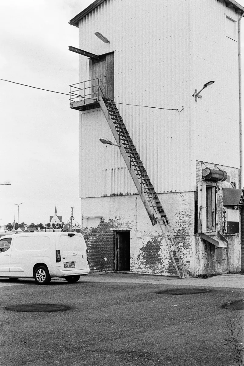 B&W industrial scene showing a white corrugated building with a steep exterior metal staircase and a parked Peugeot van.