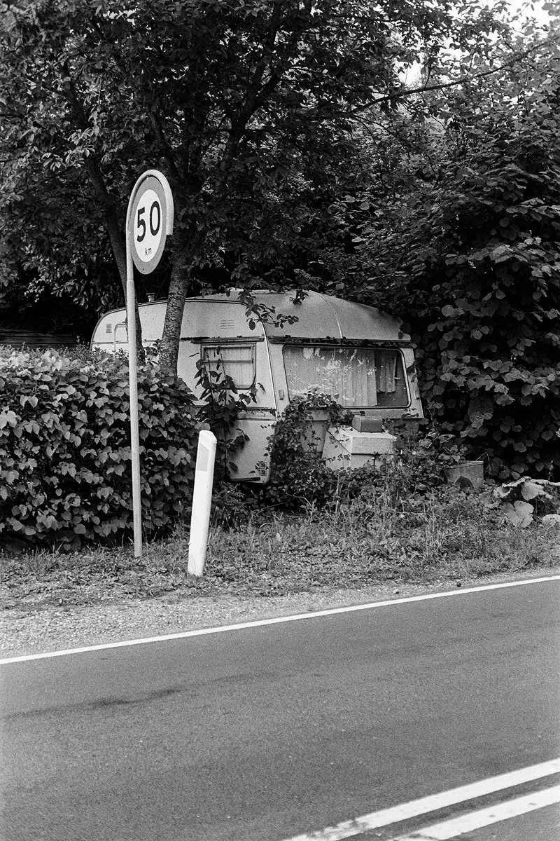 Grainy B&W shot of an abandoned vintage caravan overgrown with thick ivy behind a 50 km/h speed limit sign by a road.