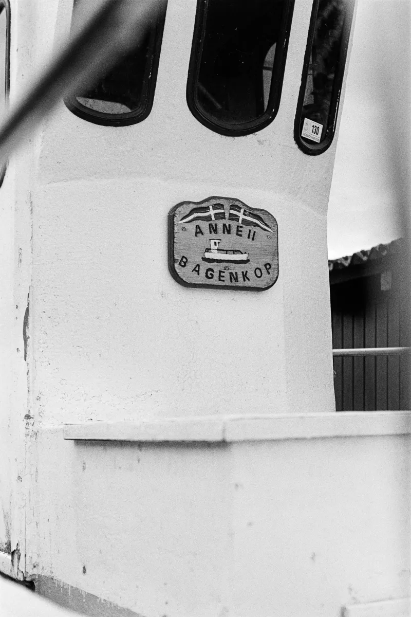 Close-up B&W photo of a white ship's bridge with three windows and a wooden nameplate for "ANNE II BAGENKOP" from Denmark.