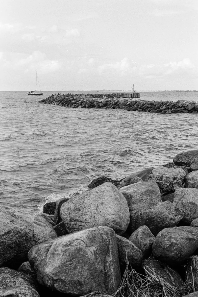 B&W coastal landscape featuring a long stone jetty, a distant sailboat, and large boulders in the foreground.