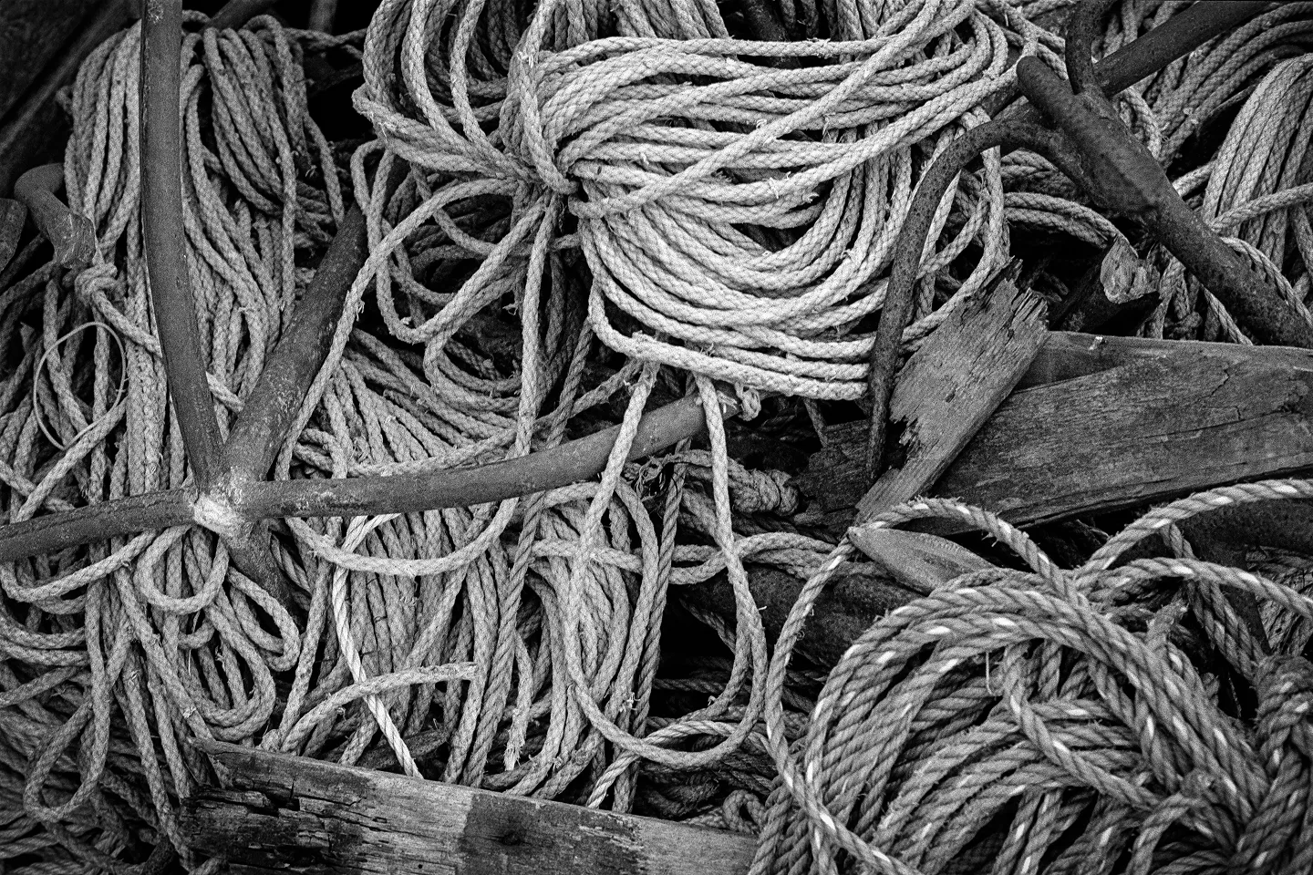 B&W abstract texture of tangled nautical ropes, heavy iron anchors, and weathered wood scraps in a high-contrast pile.