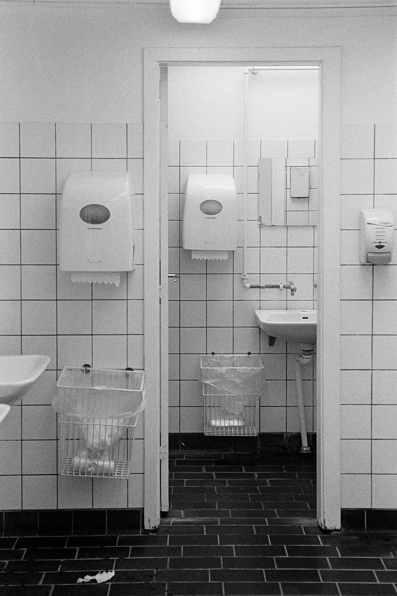 B&W symmetrical view of a public restroom with tiled walls, dual paper towel dispensers, wire trash bins, and a doorway.