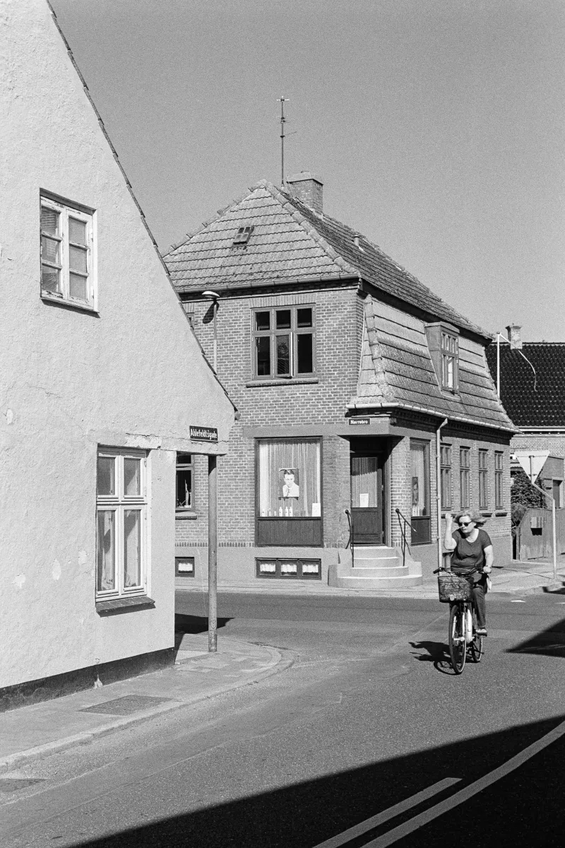 B&W street scene of a woman riding a bicycle past a traditional brick building on a sunny corner in a Danish town.