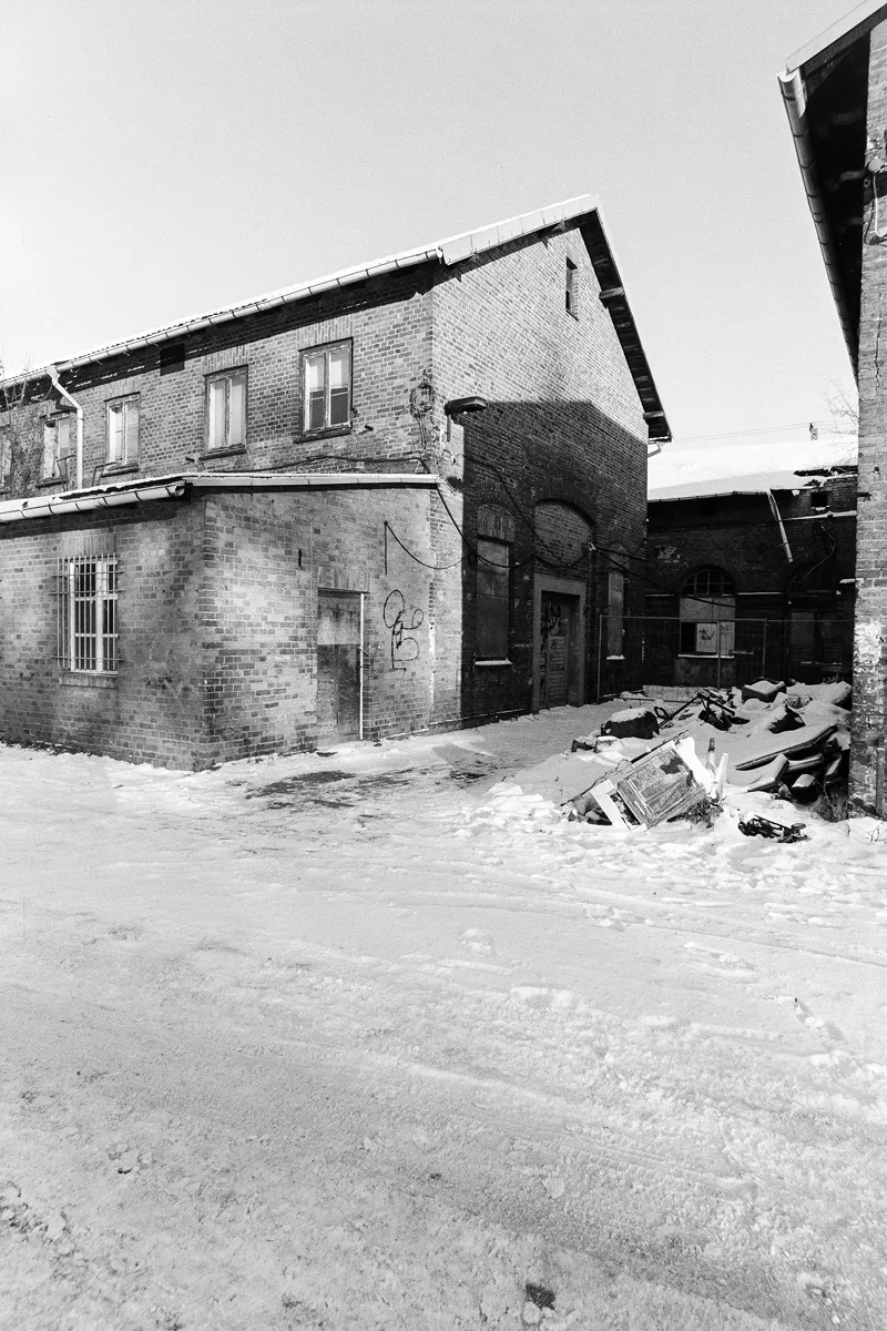 Grainy B&W shot of a weathered brick building in a snowy yard with piles of debris and industrial winter textures.