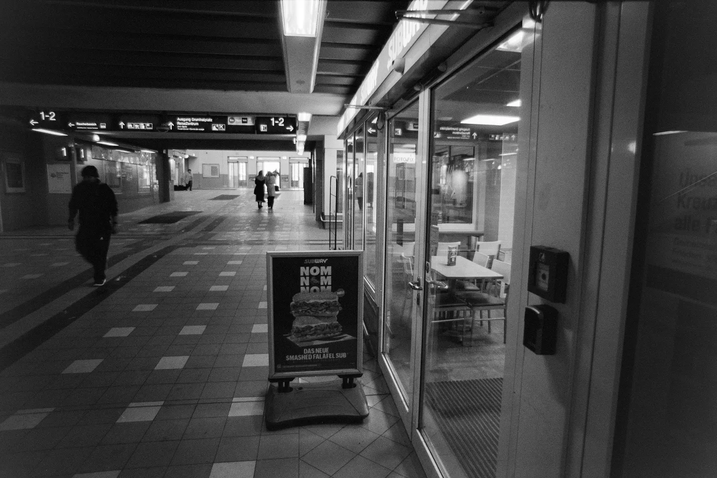 B&W film photo of a dimly lit train station interior featuring a Subway restaurant and a "Nom Nom Nom" advertising poster.