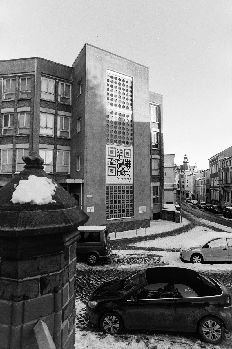 B&W film photo of a DDR-era modernist building in Schwerin with a large QR code and geometric facade on a snowy street.