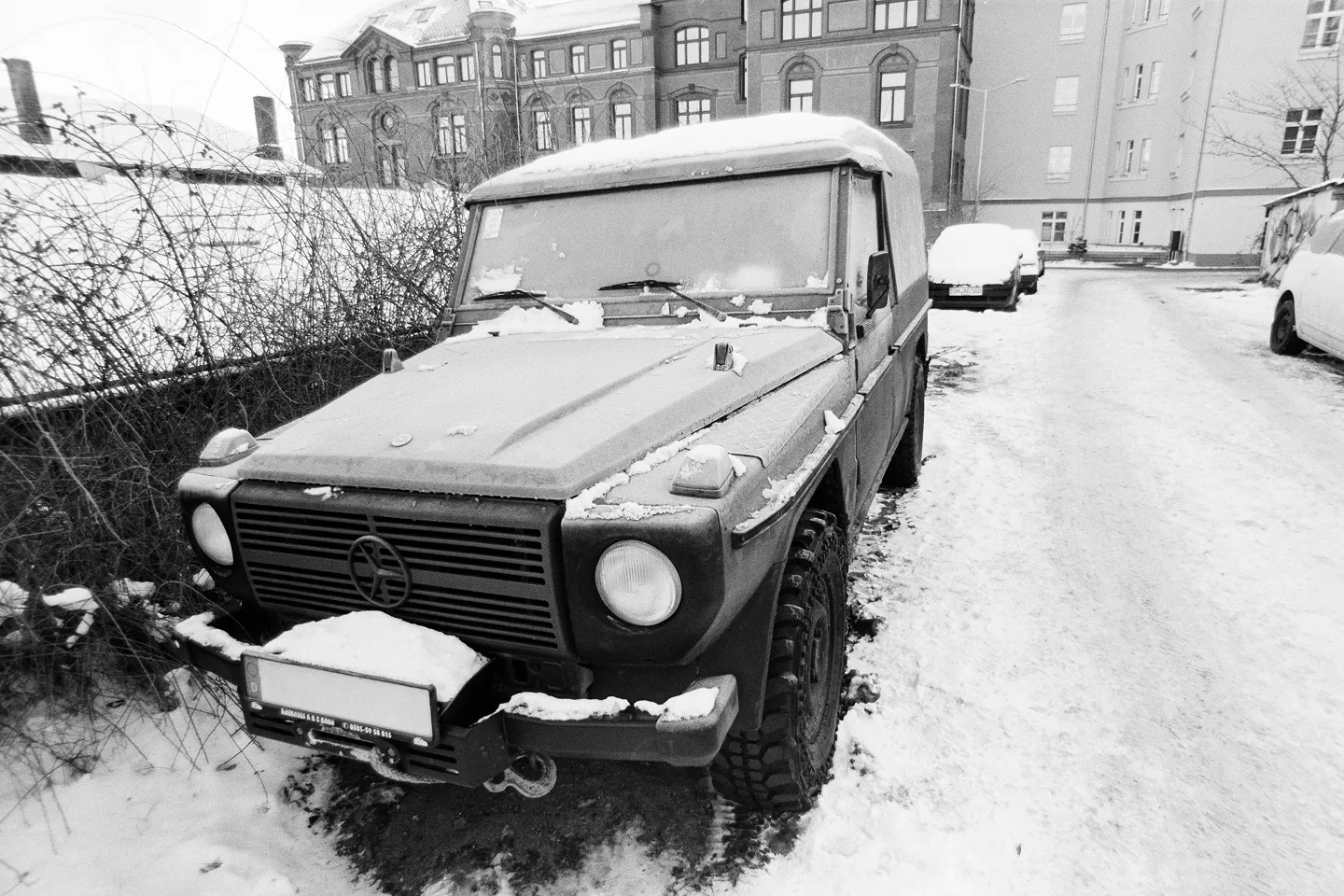 Grainy B&W shot of a snow-covered G-Wagon on a winter path with tangled bushes and historic buildings in the background.