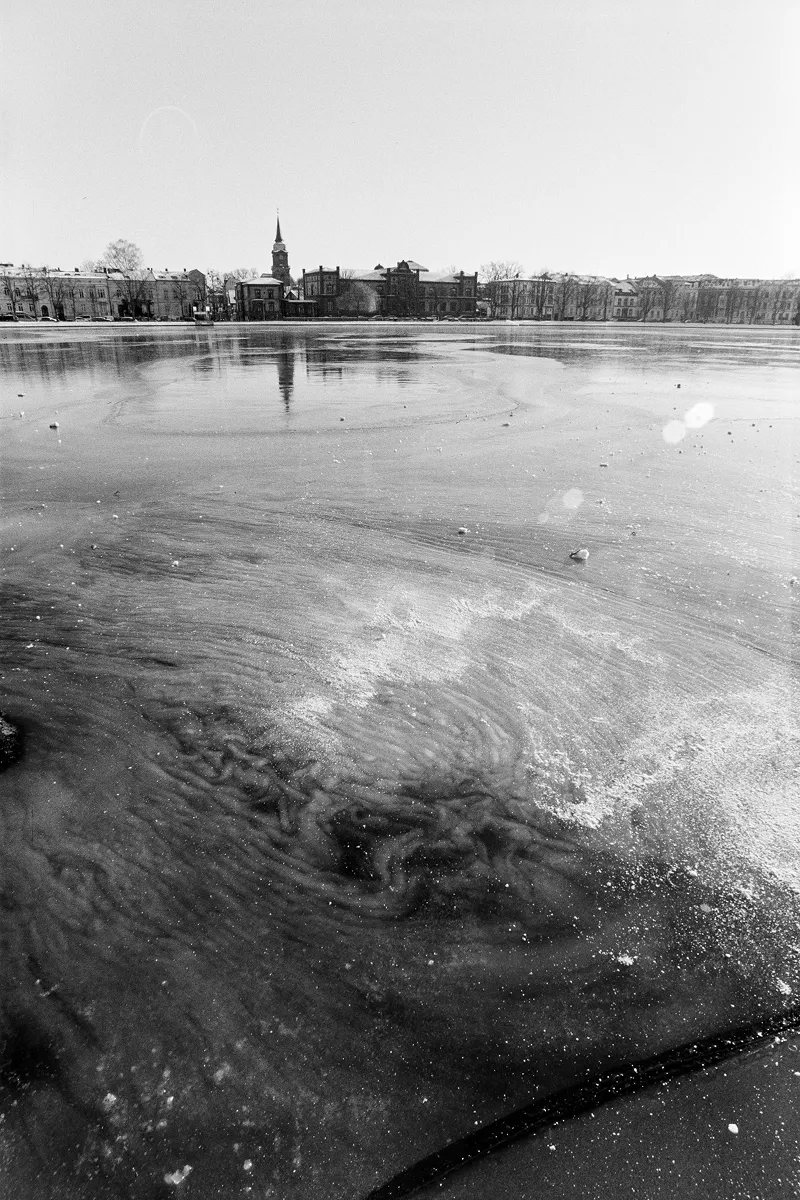 Wide B&W film photo of a frozen lake with swirling ice patterns and the Schwerin city skyline in the far distance.