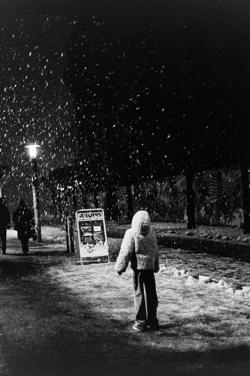 Grainy night photo of a child in a white hooded jacket standing in a snowfall under a glowing streetlamp.