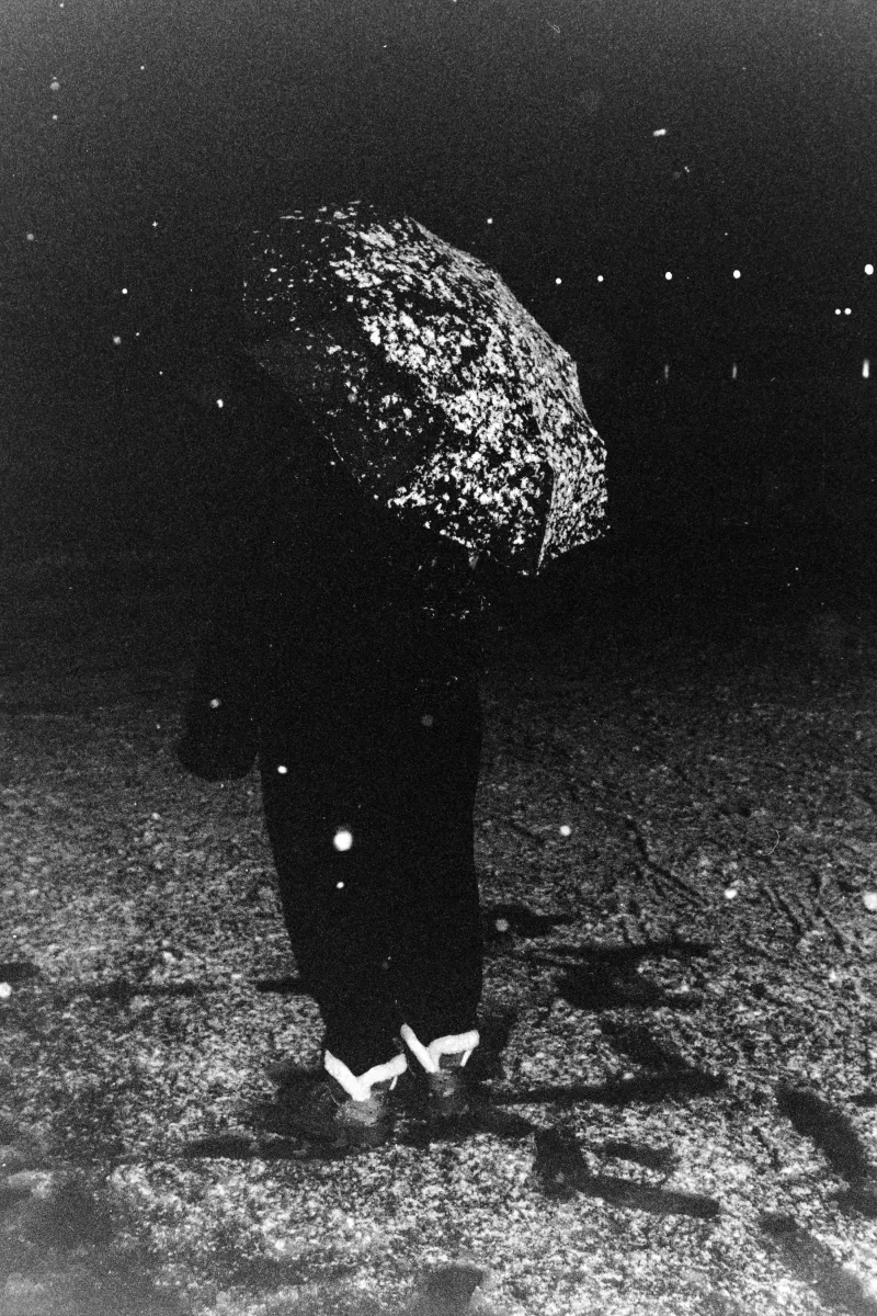 Dark, grainy B&W shot of a person from behind holding a snow-covered umbrella during a blizzard in the late evening.