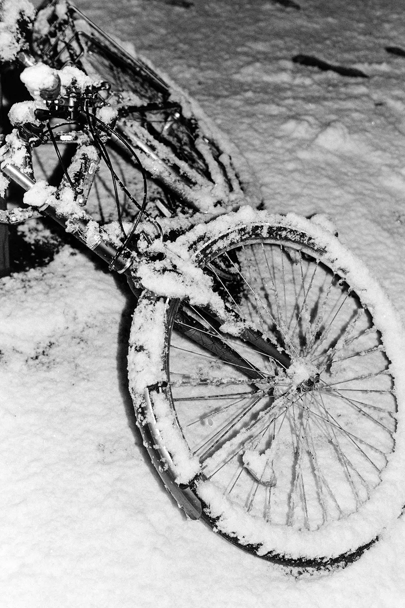 Close-up B&W photo of a bicycle lying on the ground, heavily covered in a thick layer of fresh, white snow.