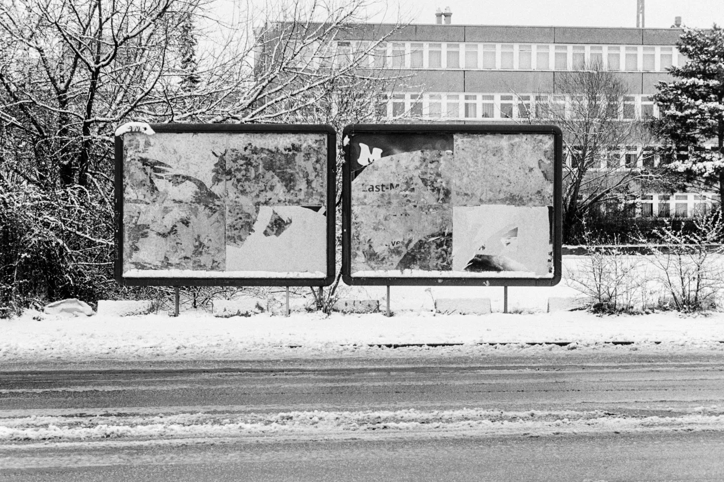 Two empty, weathered billboards in a snowy field with a building and snow-covered trees in the background.