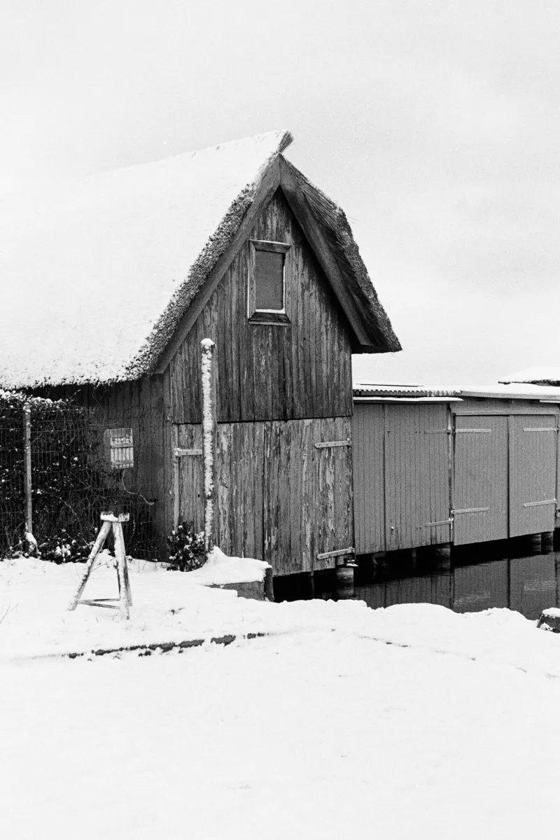 B&W film photo of a rustic wooden boathouse with a thatched roof in a snowy landscape. High contrast and weathered textures.
