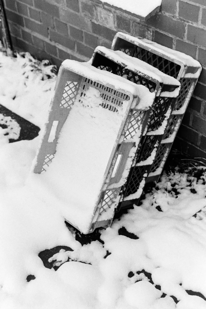 B&W film photo of stacked plastic crates leaning against a brick wall, partially buried in textured, grainy snow.