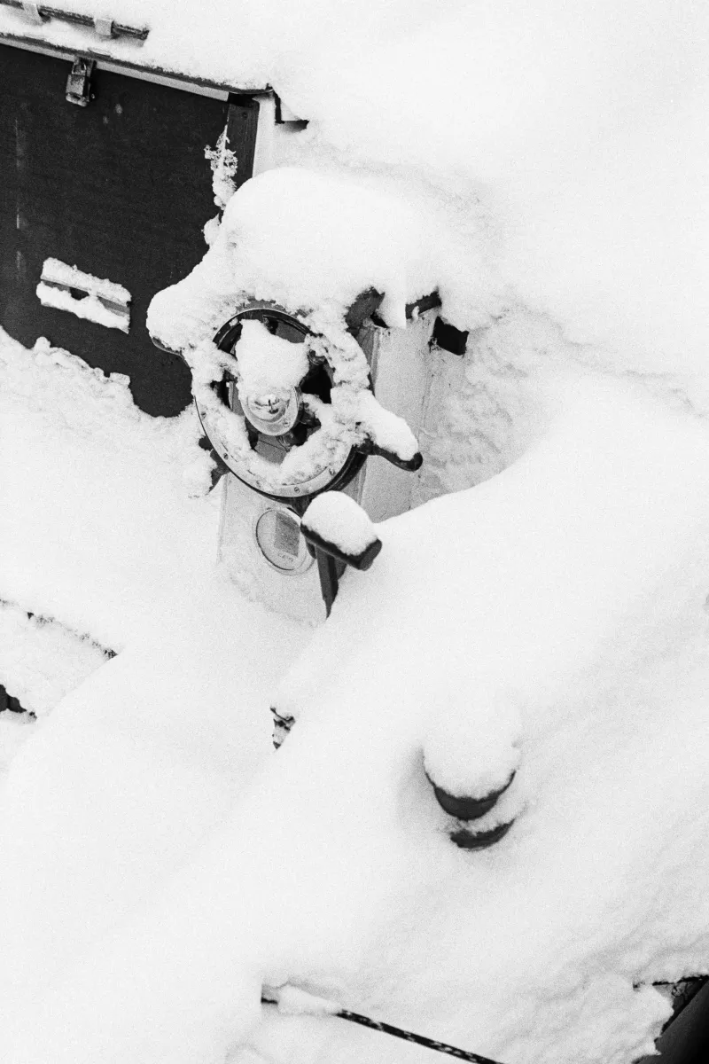 Grainy B&W close-up of a ship's steering wheel and throttle heavily blanketed in thick, fresh winter snow.