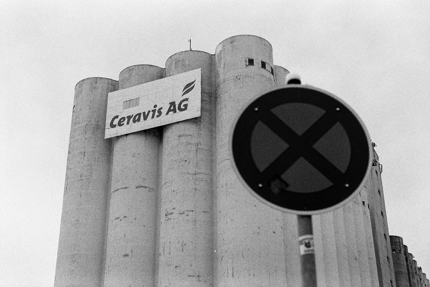 Grainy B&W photo of Ceravis AG industrial silos in Schwerin with a blurred traffic sign in the foreground.
