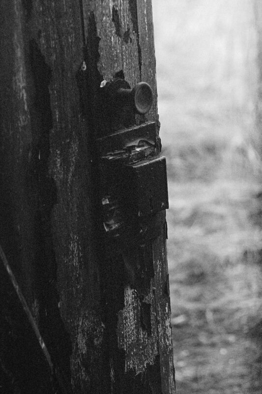 Grainy B&W close-up of a rustic metal latch and handle on a textured, peeling wooden door with high contrast shadows.