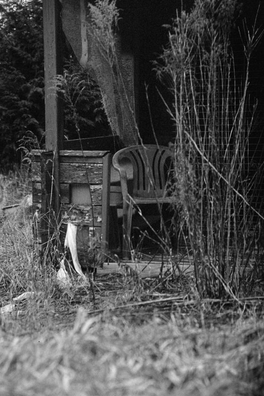 B&W shot of a plastic chair on a cluttered porch, partially obscured by tall dry weeds and a hanging mesh fabric.