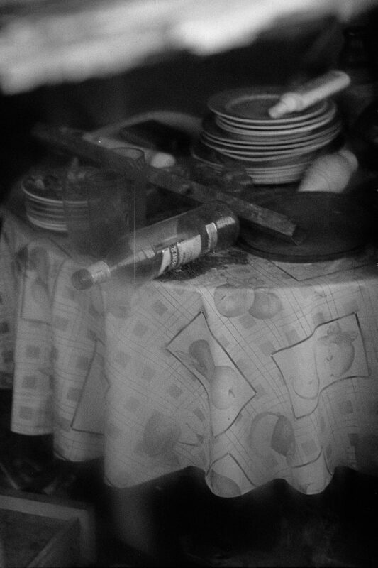 Grainy B&W photo of a cluttered table with a patterned cloth, featuring stacked plates and a glass bottle lying on its side.