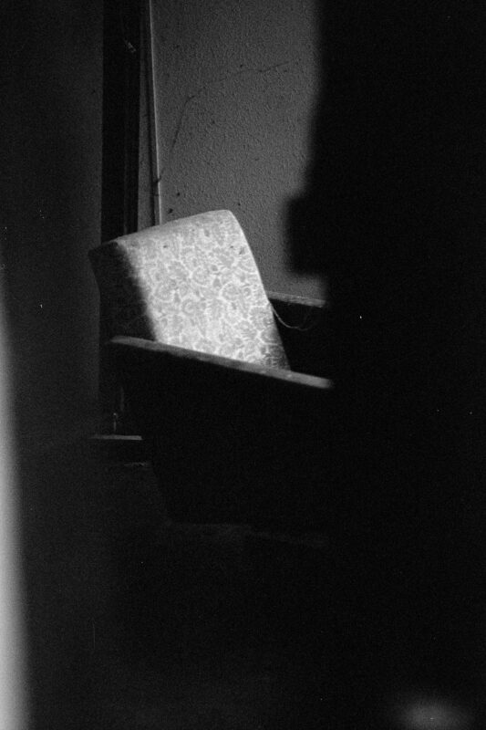 B&W film photo of a patterned armchair in a dark interior, illuminated by a sliver of light against a textured wall.