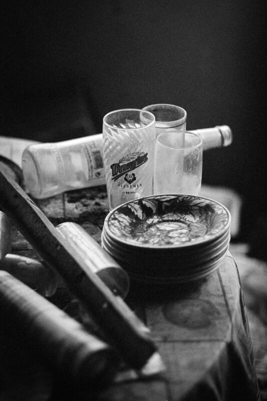 B&W close-up of a Wernesgrüner beer glass and stacked plates on a table, with a discarded bottle in the background.