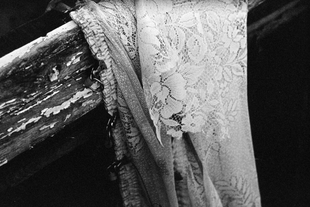 Grainy B&W film photo of a tattered lace curtain hanging from a missing window frame in a derelict structure.