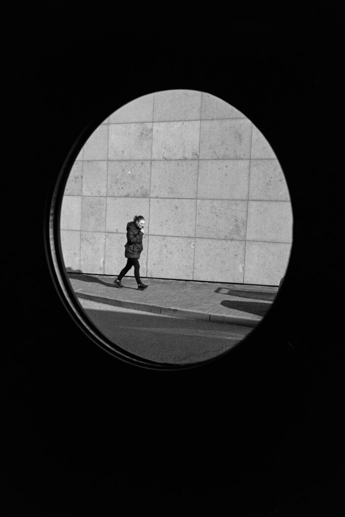 B&W film photo of a person walking on a sidewalk, framed by a dark circular porthole, grainy textures.