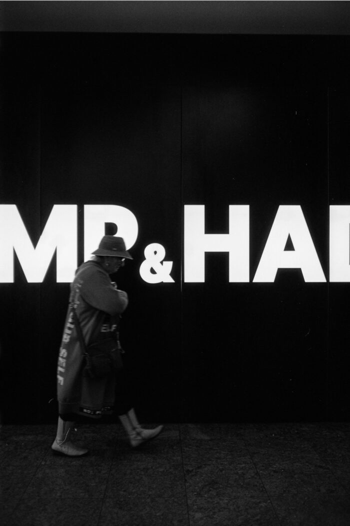 Grainy B&W shot of a hooded figure walking past the large illuminated sign text 'MP & HAI'.