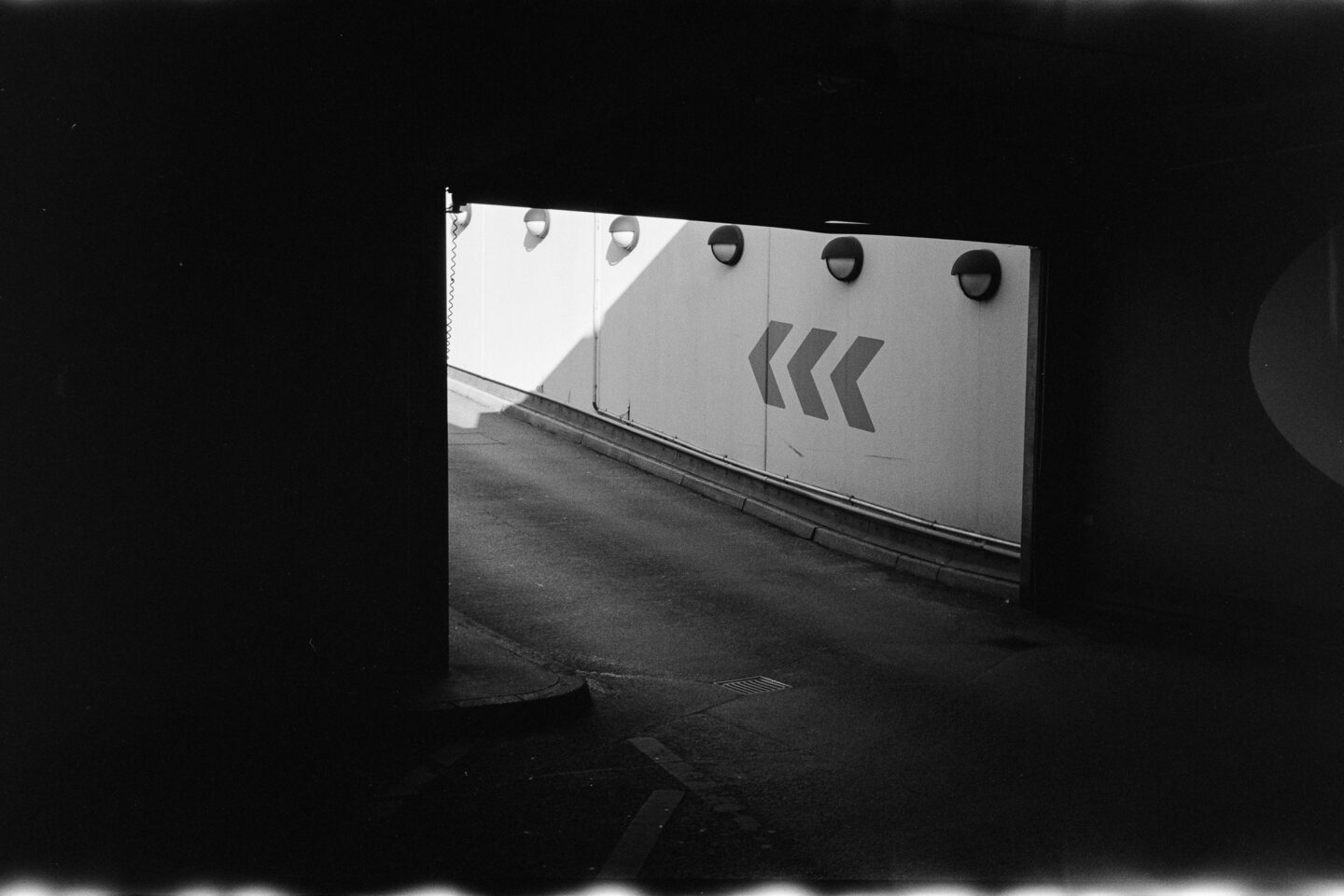 B&W film photo looking out from the shadows into a lit parking garage entrance ramp with arrows.