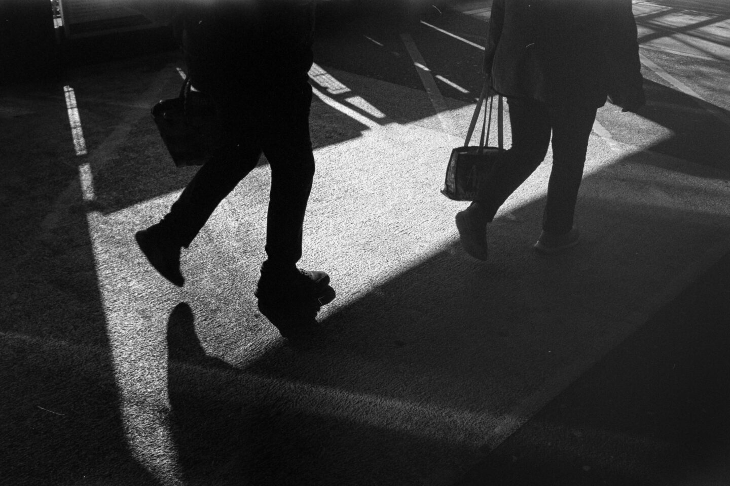 Close-up B&W film photo of two pairs of legs and their long shadows walking on weathered concrete.