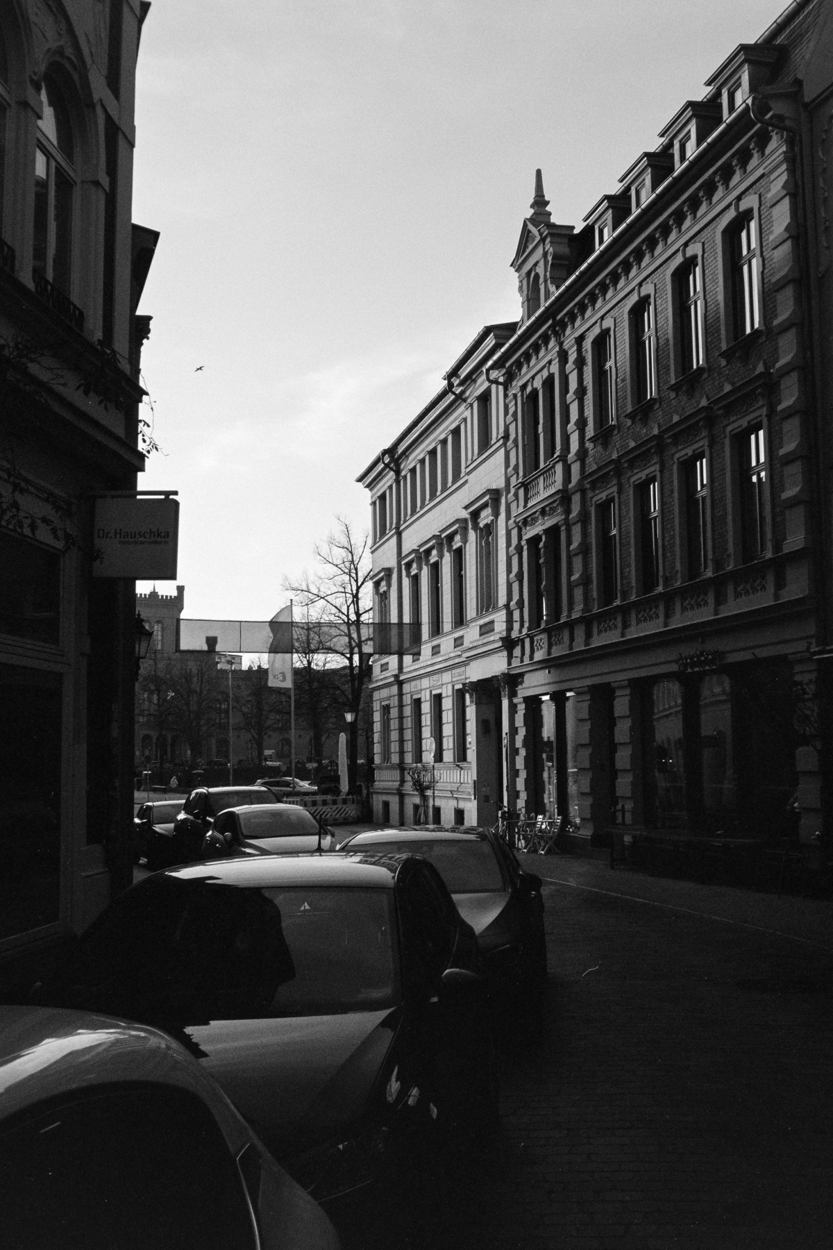B&W film photo of a street in Schwerin with parked cars and historic facades under a bright sky. Grainy textures.