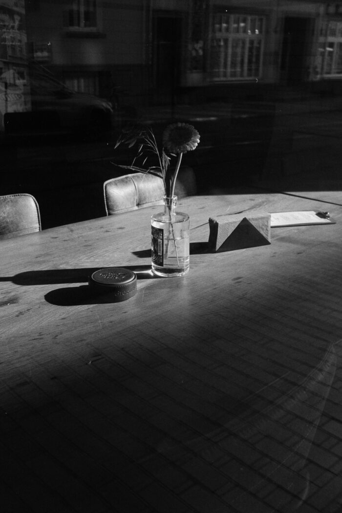 Grainy B&W film photo of a single flower on a sunlit cafe table with sharp shadows and city reflections in the window glass.
