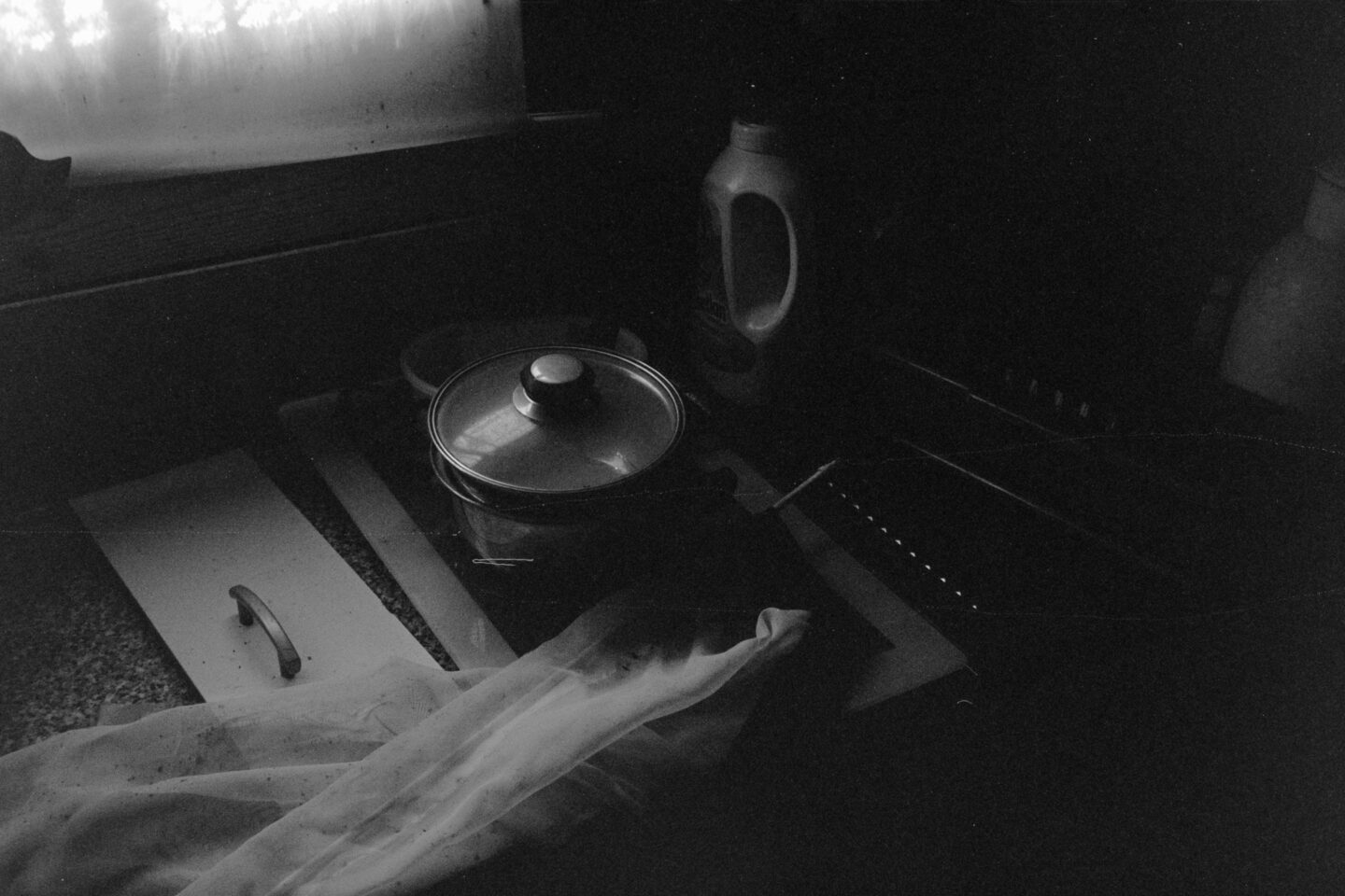 Gritty B&W film photo of a kitchen stove with a pot, captured in deep shadows with heavy film grain textures.