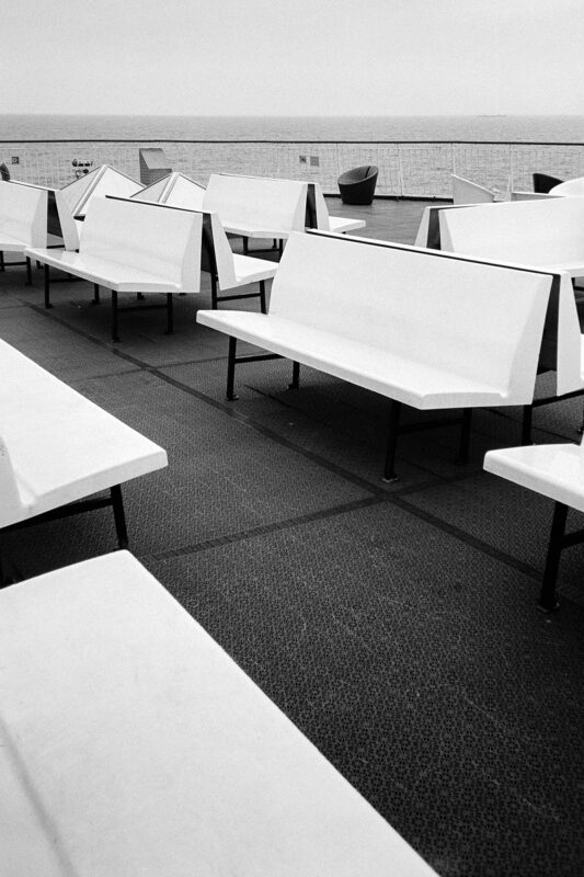 B&W film photo of geometric white benches on a ferry deck overlooking a calm sea. Grainy, minimalist composition.