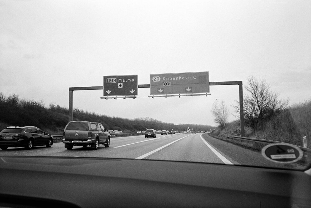 B&W film photo of highway signs for Malmø and København. Grainy travel shot from inside a car on a Danish motorway.