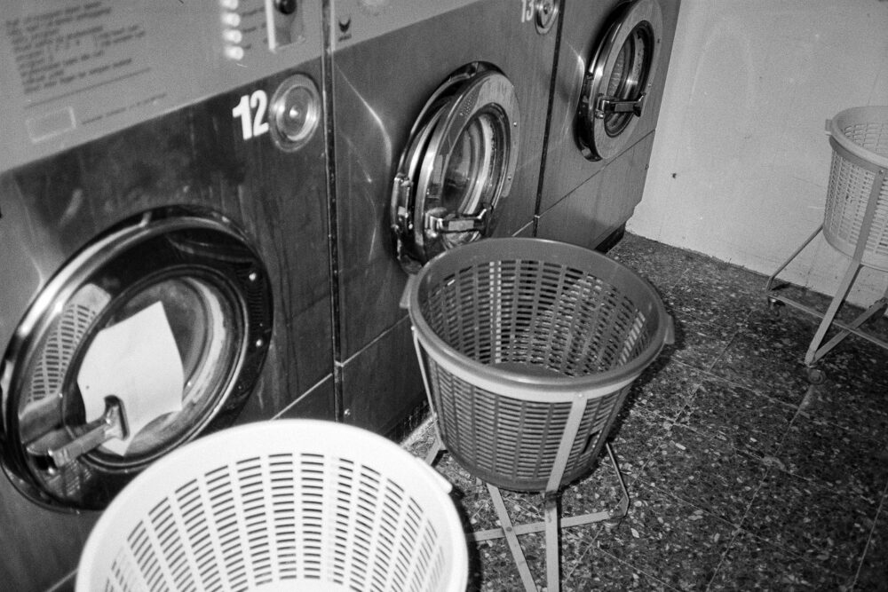 B&W film photo of industrial washing machines and plastic laundry baskets. High contrast, grainy textures in a laundromat.