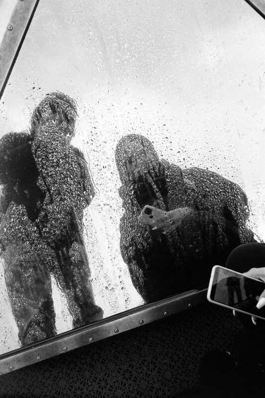 B&W film photo of silhouettes reflected in a window covered in raindrops. Moody, grainy texture with abstract shapes.