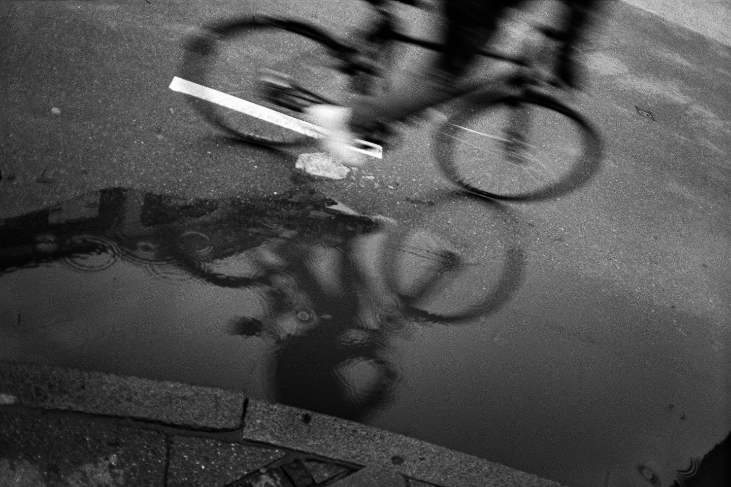 B&W film photo of a blurred bicycle passing a puddle on pavement. Dynamic motion and grainy textures in the rain.