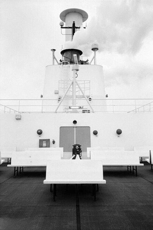 B&W film photo of symmetrical white benches and a smoking ship funnel. Minimalist, clean lines with a grainy analog look.